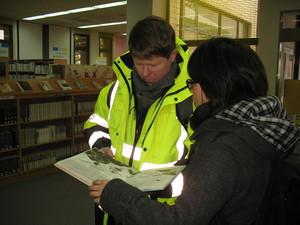 本棚が並んでいる図書館で開いた絵本を手に持って見ているキャメロンさんと日本人女性の写真