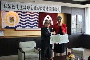 「姉妹港支援調印式並びに姉妹校締結式」と書かれた横断幕と、リズモー市の旗、大和高田市の旗が掲げられた室内で、吉田市長とジェニー・ドウェル市長が同じ書類をそれぞれ持ち握手をしている写真