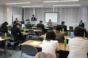 並べられた長机に参加者達が座り、前方に置かれたスクリーンの横に男性が立ち定期総会が行われている写真
