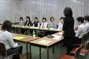 四角に並べられた机にたくさんの人達が座り一人の女性が立っている写真