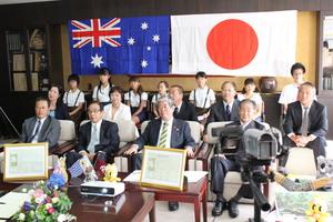日本の国旗とオーストラリアの国旗が並ぶ部屋で式典に参加しているたくさんの人達が椅子に並んで座り記念撮影をしている写真