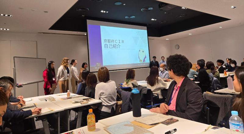 テーブルで座り、会議室の前の画面を向いている大人数の様子。画面の横に、6人の発表者が立っている様子。