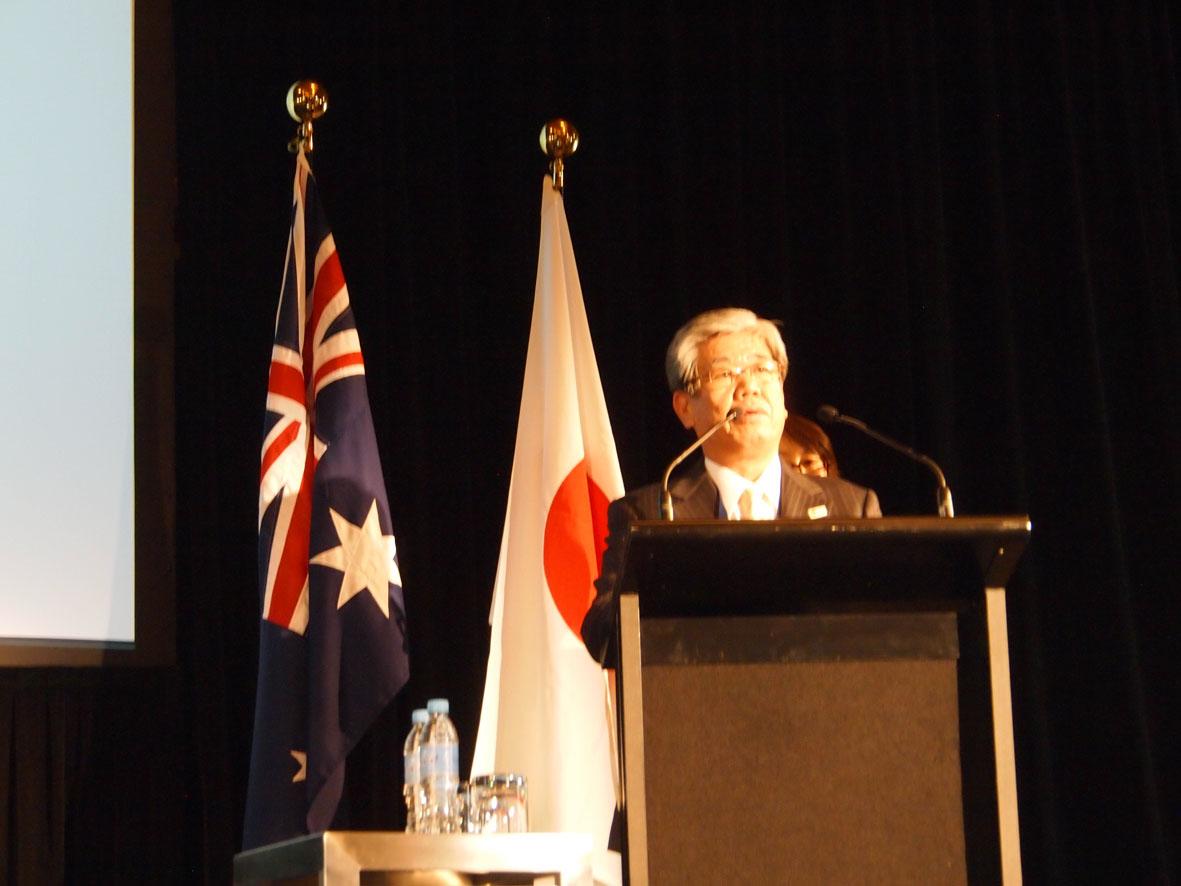 オーストラリアと日本の国旗が飾ってある舞台で挨拶をしている男性の写真