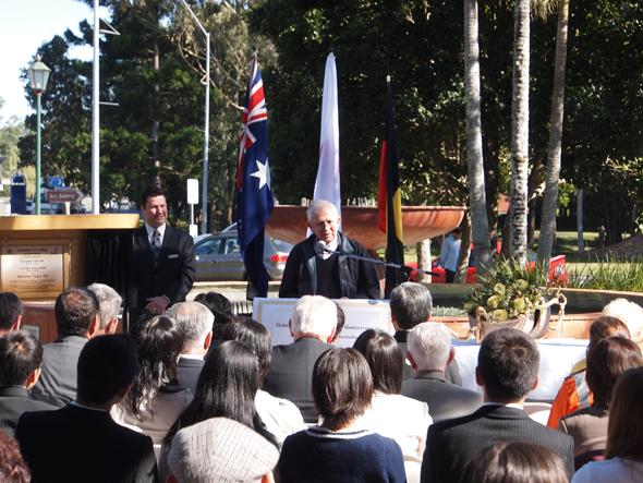 オーストラリアと日本の国旗が飾られたセレモニーで挨拶をしている白髪の男性の写真