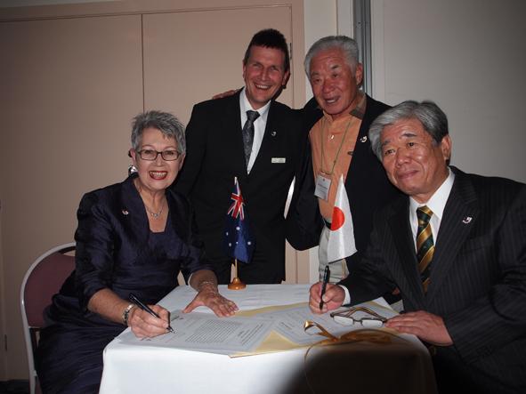 小さなオーストラリアと日本の国旗が飾られたテーブルの周りに4名の人が笑顔で並び記念撮影を撮っている写真