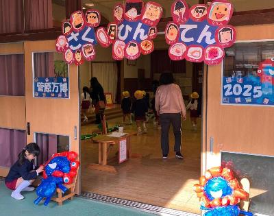 教室の入り口で二つのみゃくみゃくの風船が開いているドアの左右に置かれ、1人の女の子が左の風船を直している様子。教室の中で展示の作品を見つめている人の様子