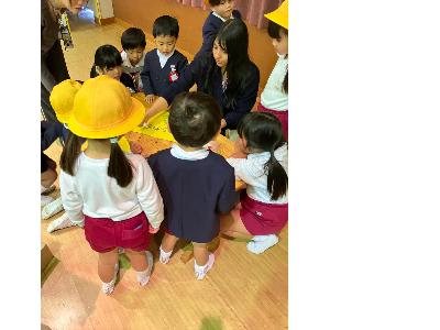 テーブルに囲って8人の園児たちが、文字を書いている女性を見つめている様子