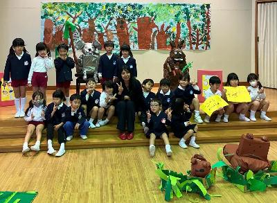 前に向き、1人の女性と大人数の園児たちは浅いステージに座り、写真のの様子。周囲に作品がある。