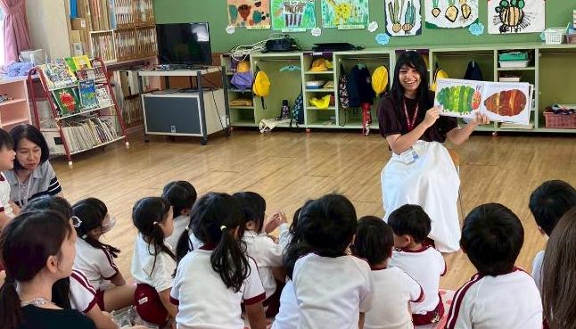 大勢の座っている園児たちの前で、絵本を手に持って読み聞かせをしている女性の様子。