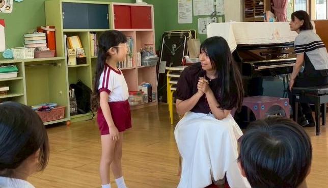 幼稚園の部屋で、1人の女性が女の子の園児に話しかけている様子。