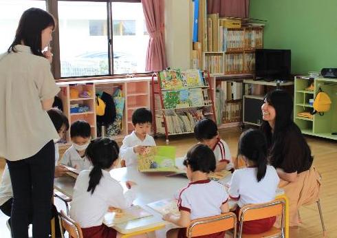 教室の中で、子供達と1人の女性がテーブルで座っている様子。子供達は本を読んでいる様子。
