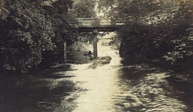 両側に木々が生え、中央に川が流れている奥に橋が見える昔の白黒写真