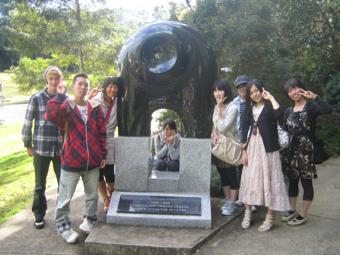 1人の外国人の男性と7名の日本人学生が黒いモニュメントを囲みピースサインをして記念撮影をしている写真