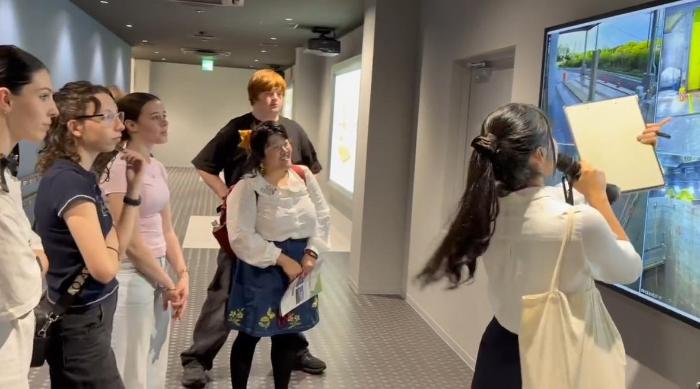 5人の人の前で1人の女性がテレビの画面に向けて、説明している様子