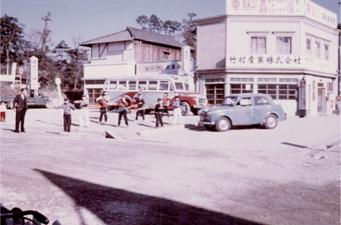 レトロな建物の前に昔の車とバスの横に人々が立っている白黒写真