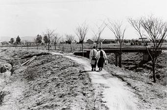 堤防のあぜ道を歩く女性2人の後ろ姿を写した白黒写真