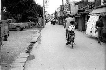 お店の前を自転車で2人乗りをしている人を後ろから写した白黒写真