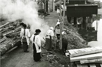 かっぽう着姿の女性たちが煙の周りに立っている白黒写真