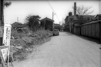 左手前に病院入り口と書かれた看板が立ち、道路を挟んだ奥に高い煙突の建物が写された白黒写真