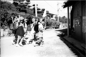 舗装されていない道路を集団で手を挙げて歩いている小学生の子供たちの白黒写真