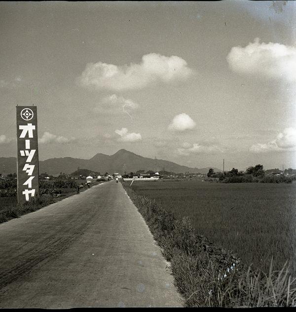 左道路脇にオーツタイヤと書かれた看板があるのどかな田園風景の白黒写真