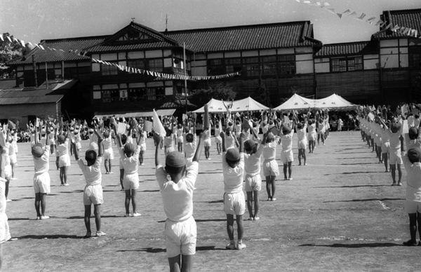 木造の校舎の前の運動場で白色の上下の体操服をきた児童が両手を上にあげ体操をしている白黒の運動会の写真