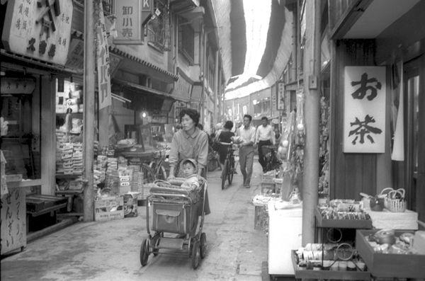 商店街のアーケード内を乳母車に子供を乗せ買い物している人や自転車を押して歩いている人などの白黒写真