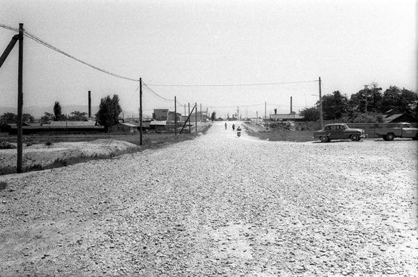 舗装されていない広い道路の両脇に電信柱がたち、車が停車している白黒写真