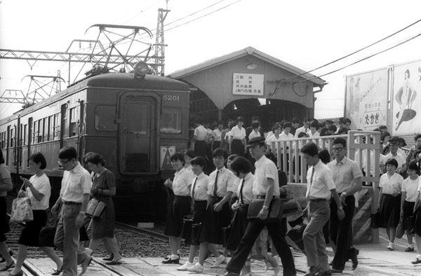 たくさんの人達が駅構内から下車し電車が停車している踏切を渡っている白黒写真