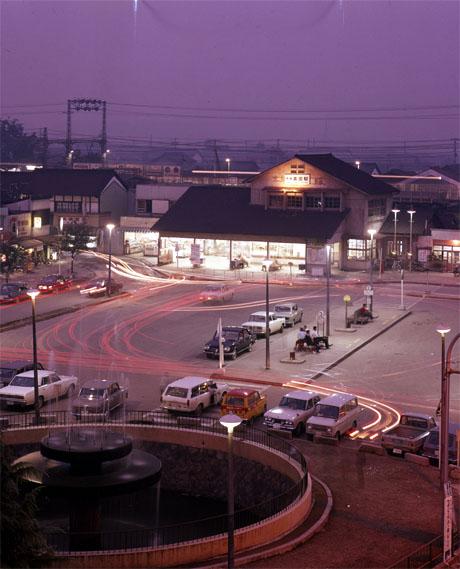 近鉄大和高田駅舎とその前の道路に停車する車、バス停で待つ人たちを空撮した夜景の写真