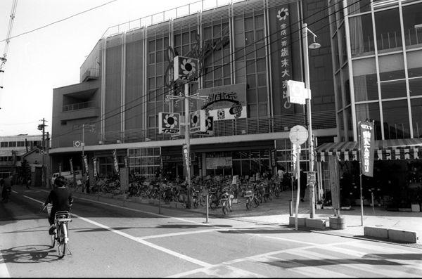 右側にスーパーいずみやの店舗があり、その前の道路を自転車で通行している人を撮影した白黒写真