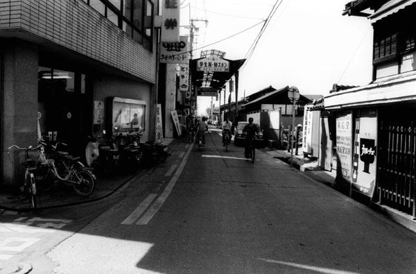 道路両脇に店舗が並ぶ商店街入り口の一方通行の道路を写した白黒写真