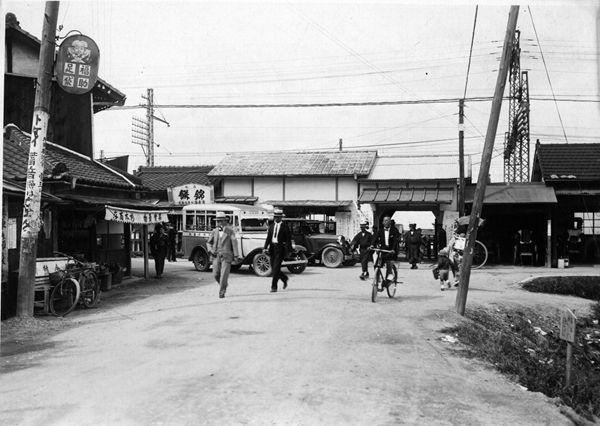 道路には車が停車、帽子を被った男性が歩き、自転車に乗っている人がいる駅前の白黒写真