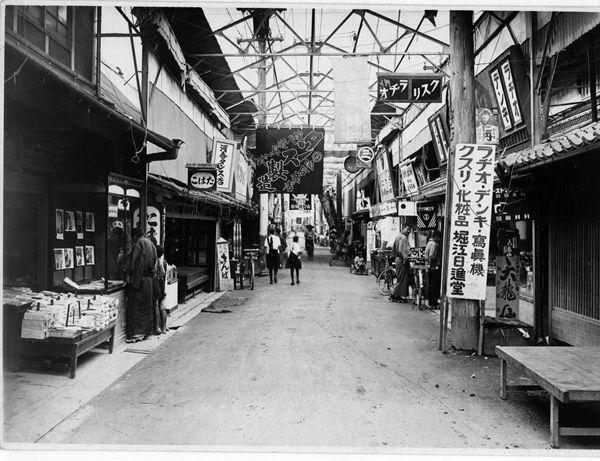 商店街のアーケード内の店舗で買い物をしている人の白黒写真