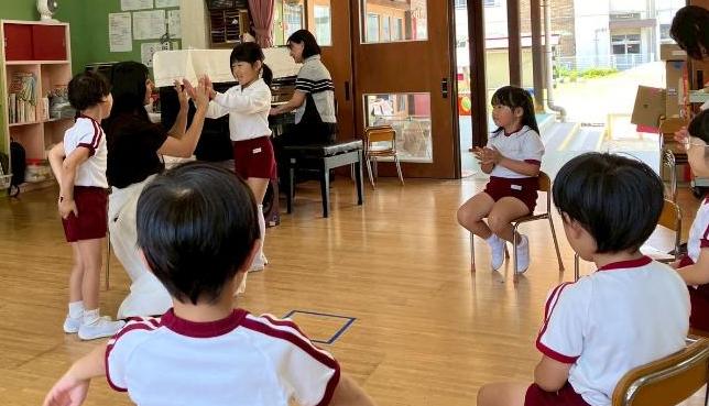 幼稚園の部屋で、1人の女性が女の子の園児とハイタッチをしていて、周りでは5人の園児たちがその様子を見ている。