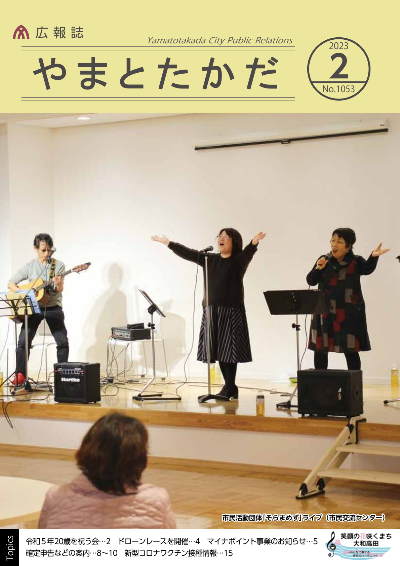 広報誌2月号表紙  コスモスプラザにて、市民活動団体である「そらまめず」の三人が音楽ライブを行っている様子（左にギター演奏者が一人、真ん中と右に手を広げて歌っているボーカル二人）