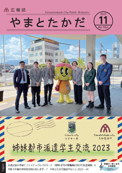 広報誌11月号表紙  大和高田市の姉妹都市であるリズモー市からの派遣学生5人と、随行者1人、大和高田市のマスコットキャラクターみくちゃんの写真。庁舎前でみんながこちらを向いて微笑んでいる様子。