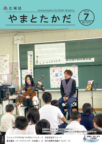 広報誌7月号表紙 市内の小学校にて、カウンターテナー歌手の藤木大地さんとクラシックギタリストの猪井亜美さんがコンサートを行っている様子。二人とも椅子に座り、猪井さんの伴奏に合わせて藤木さんが歌っており、それをたくさんの児童たちが見ています。